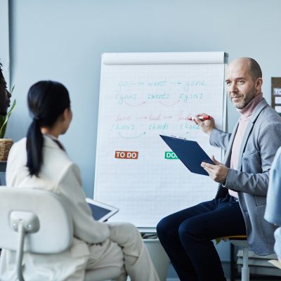 Portrait of smiling mature coach giving English lesson to group of people sitting in circle during seminar in office